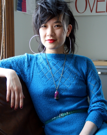 An Asian woman with rad hair wears a blue knit dress and giant lilac hoop earrings.